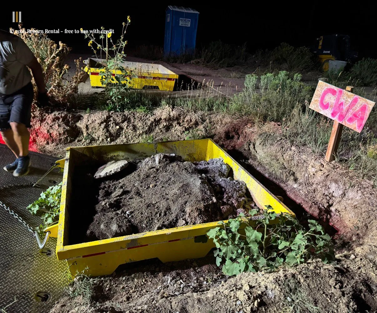Chains winching a yellow steel concrete washout pan filled with concrete slurry onto a flatbed truck during an evening pickup operation.