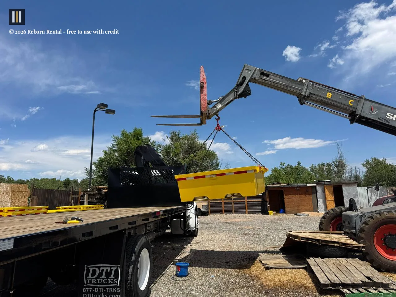A telehandler lifts a yellow concrete washout pan with chains from the bed of a flatbed truck